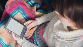 A woman wearing a white sweater and covered with a colorful blanket holding a tablet in her hand and...