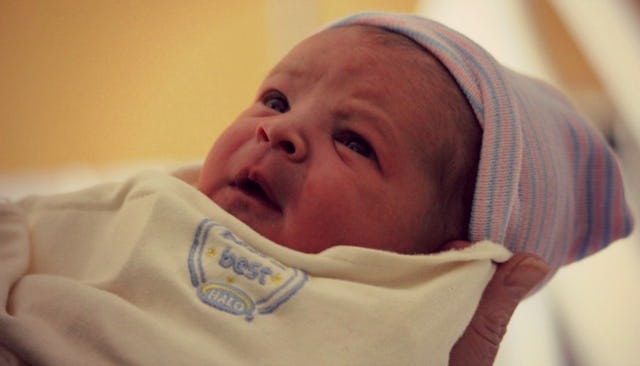 A newborn baby in his mother's arms, wrapped in a yellow blanket and a purple-and-blue striped cap.