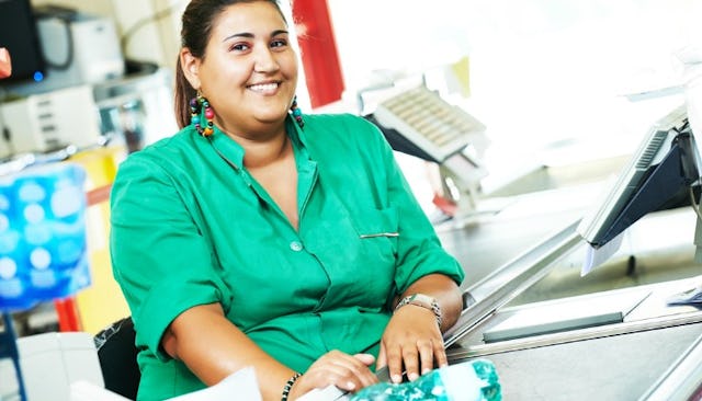 A retail worker at the cash register, dressed in a green uniform with beaded hoop earrings and hair ...