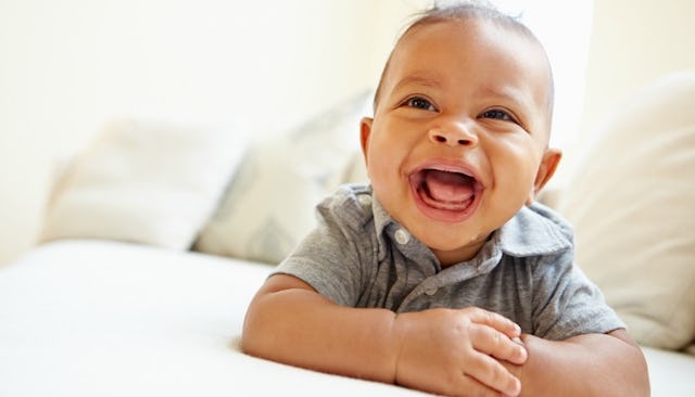 A toddler lying on his stomach on a beige couch wearing a gray T-shirt with crossed arms laughing