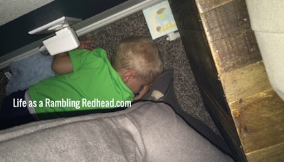 A child in a green T-shirt sleeping on the floor behind a piece of furniture 