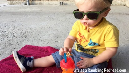 A child with sunglasses eating fast food while sitting on a burgundy towel that’s on the ground