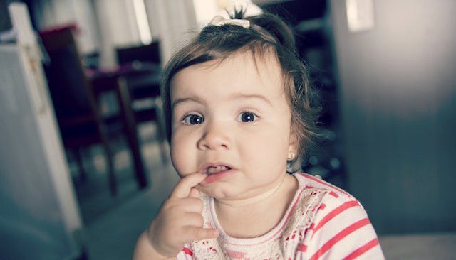 A one-year-old girl holding her index finger inside her mouth while wearing a pink and white shirt