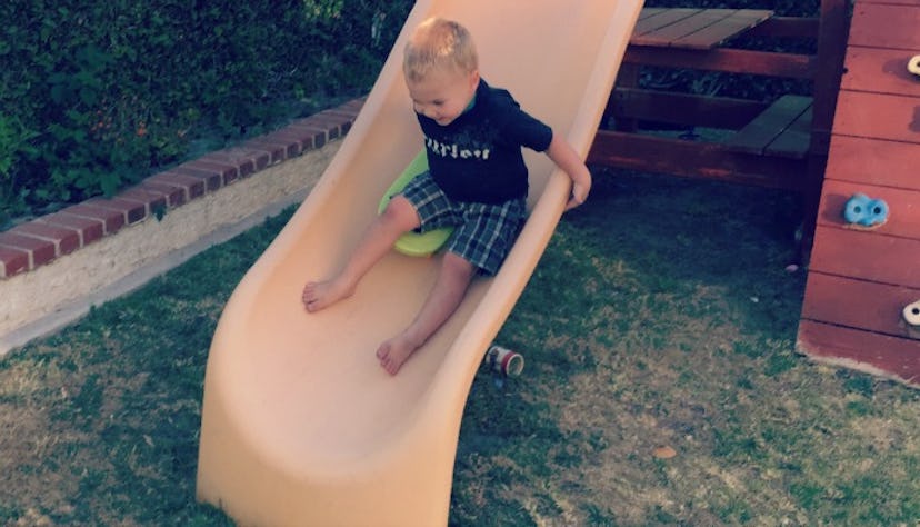 An autistic blonde boy in a black T-shirt and green checked shorts laughing on a yellow slide in a b...
