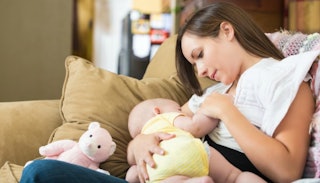 A brunette woman breastfeeding her baby in a yellow onesie on a brown couch and smiling