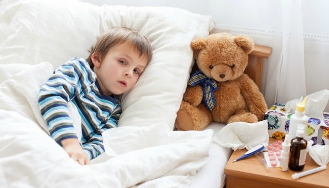 A sick kid lying in bed with a teddy bear, medicine, and tissues on the night table next to the bed