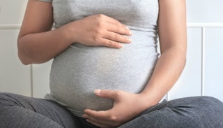 A pregnant woman holding her stomach with both hands while sitting on the floor in a grey shirt and ...