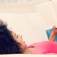 A SAHM with curly black hair and wearing a pink top is lying on abed and reading a book.