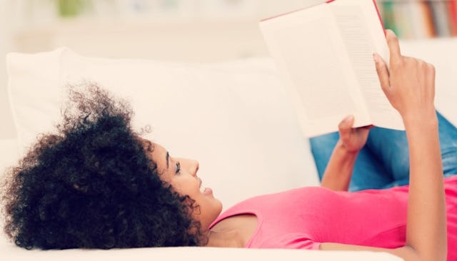A SAHM with curly black hair wearing a pink top lying on a bed while reading a book