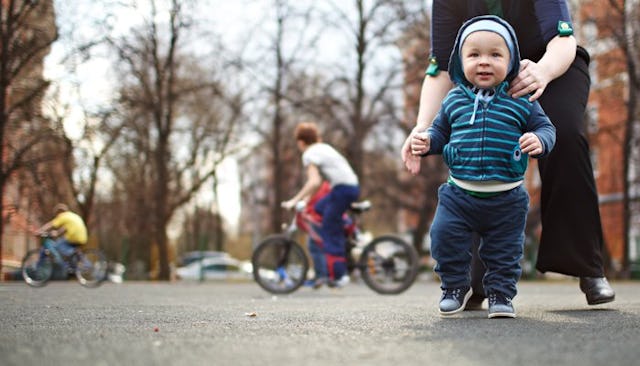 A baby making his first steps while older kids are riding bicycles in the background.