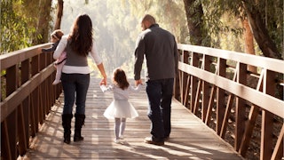 Family of four crossing the wooden bridge holding their little girl with hands and holding the baby ...