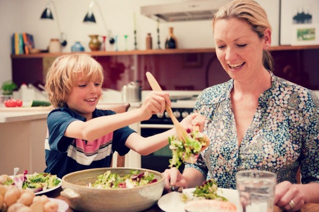 A blonde woman wearing a blouse and a blonde boy putting salad on her plate while both of them are s...