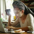 A woman with face mask, towel over her hair, and a cup of coffee using on her laptop and needs to ge...