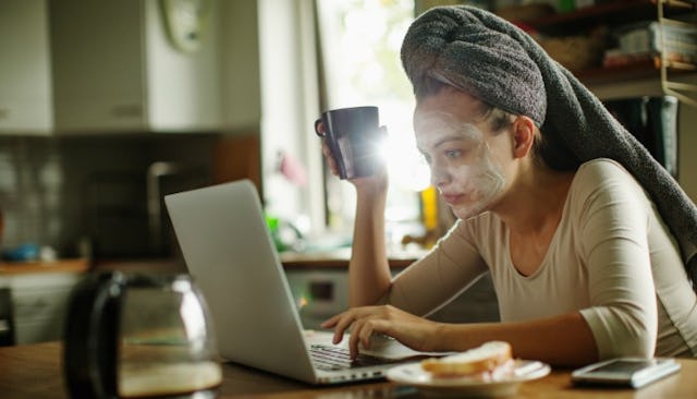 A woman with a face mask, towel over her hair, and a cup of coffee using her laptop needs to get off...