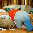 A toddler in an orange shirt grey sweatpants with his face buried in a brown pillow and a beige blan...