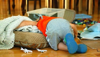 A toddler in an orange shirt grey sweatpants with his face buried in a brown pillow and a beige blan...
