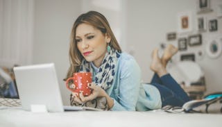 A woman lying on the bed, drinking coffee and looking at a laptop while experiencing infertility str...