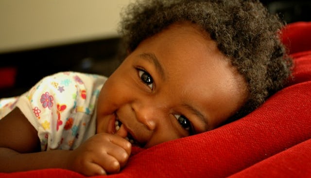 A little curly-haired baby lying down on a bed and smiling with her thumb in her mouth
