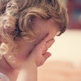 Blonde girl with curly hair holding her hands on her face like she is crying with a blurred backgrou...