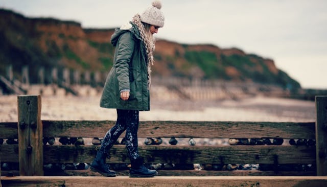A woman in her 40s walking on a wooden fence at a beach while wearing a green jacket and a white bea...