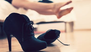 Female black high heels thrown on the floor, and a woman resting her feet in the background after he...