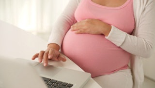 A pregnant woman in a pink T-shirt, sitting by the table working on her laptop, resting her hand on ...