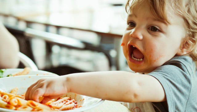 A toddler with food around his mouth, dipping his hand in a plate filled with pasta with mouth wide ...