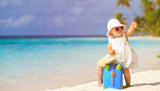 A baby sitting on a blue suitcase in the sand at the beach while holding a toy airplane.