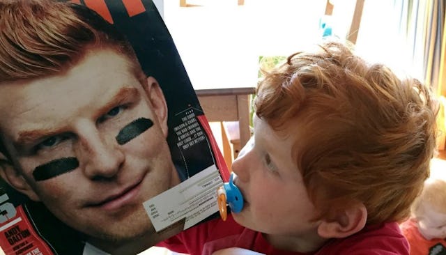 A little red-hair son in a red head shirt looking at the magazine with a red-head man on the cover o...