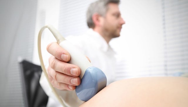 An OBGYN doctor in a white coat sitting on a black chair and holding an ultrasound scanner on a preg...