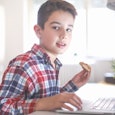 A boy at the laptop eating something and looking at the camera.