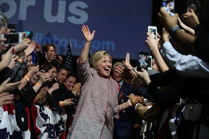 Hillary Clinton wearing a tweed pink/black/white pantsuit, waving and smiling while walking through ...