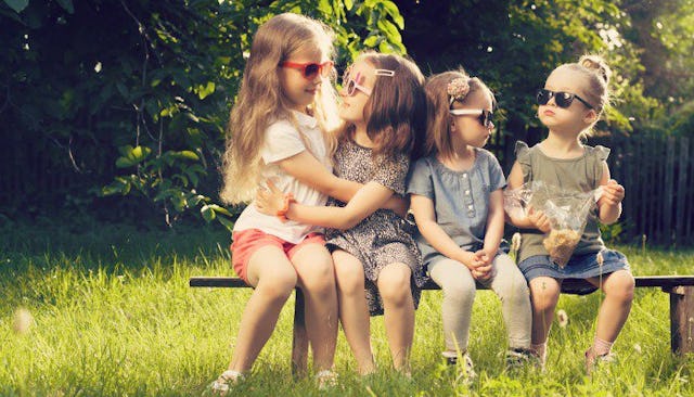 Four girls sitting on a bench in nature, all wearing sunglasses; two of them hugging each other.