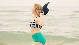 A woman with a mombod in a swimsuit holding a hat, with the sea in the background