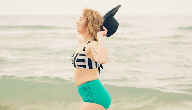A woman with a mombod in a swimsuit holding a hat, with the sea in the background