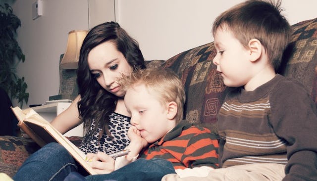 A teenage girl babysitting two boys while reading a book with them on a brown couch