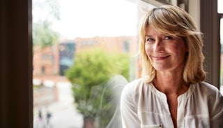A blonde woman in her 40s with a bob cut and bangs, wearing a white shirt sitting next to a window a...