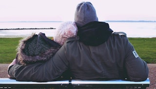A married couple sitting and hugging on a bench, looking at the sea while wearing jackets and beanie...