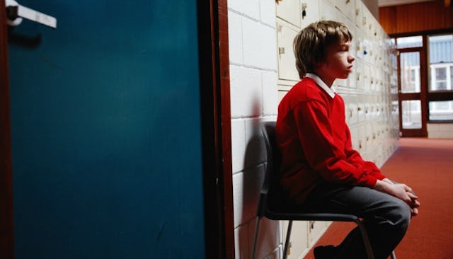 A boy with ADHD in a red sweater and black denim jeans sitting alone in a school hallway