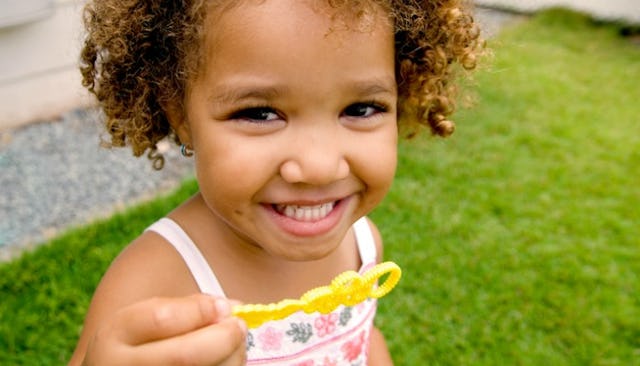 A curly-haired toddler girl standing on the grass wearing a white floral dress, smiling and holding ...