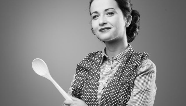 A mother in a polka dot and checkered blouse holding a wooden spoon and smiling in black and white