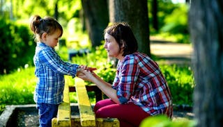 A woman and a little girl standing in nature, holding each other's hands in a park