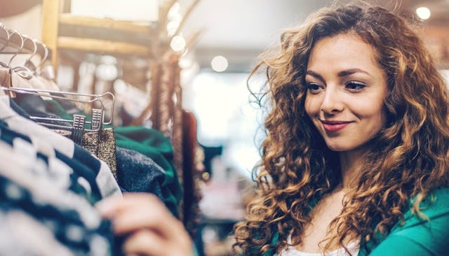 A curly hair woman in a green blouse smiling while browsing through clothes at a store, wants to buy...