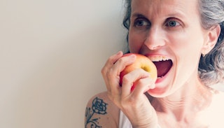 Gray-haired tattooed woman biting into an apple.