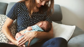 A working/breastfeeding mom with long brown hair is feeding her newborn child while she is smiling a...