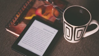 A cup, Kindle, notebook and glasses on a table.