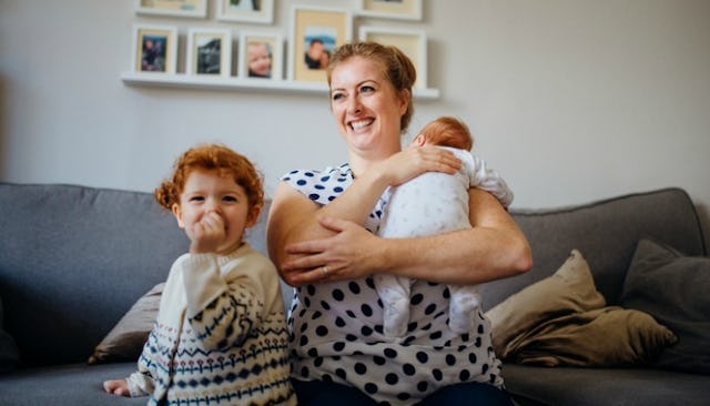 A mom sitting on a couch and smiling while holding her baby and her toddler standing next to her