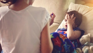 A kid with mono in bed with a stuffed bear next to him holding the hand on his face while his mother...