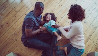 Parents tickling their kid on the wooden floor, all smiling and looking happy.