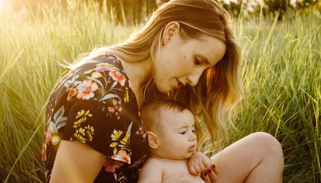 Millennial parent mom in a black floral dress sitting on grass with her newborn son